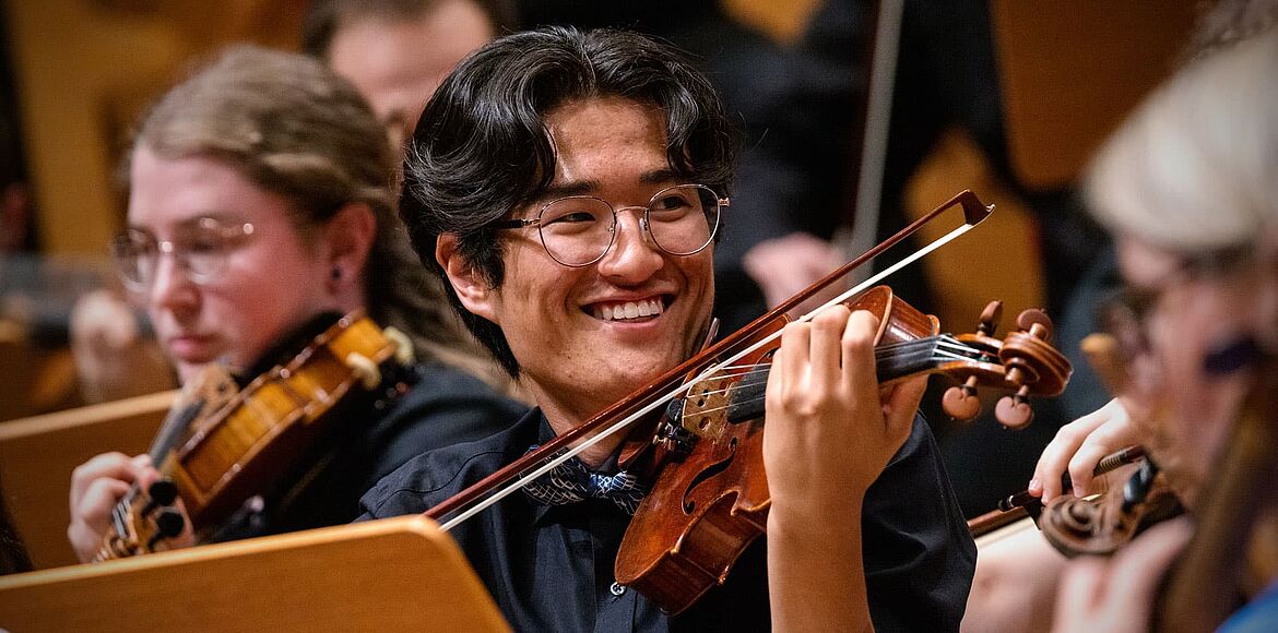 Das Bild zeigt einen jungen Geiger mit Brille, der während eines Konzerts Violine spielt und lächelnd zu einer anderen Person im Orchester blickt. Um ihn herum sitzen weitere Musikerinnen und Musiker mit Streichinstrumenten. Die warme Bühnenbeleuchtung und der enge Bildausschnitt vermitteln die konzentrierte und zugleich freudige Atmosphäre eines Orchesterauftritts.