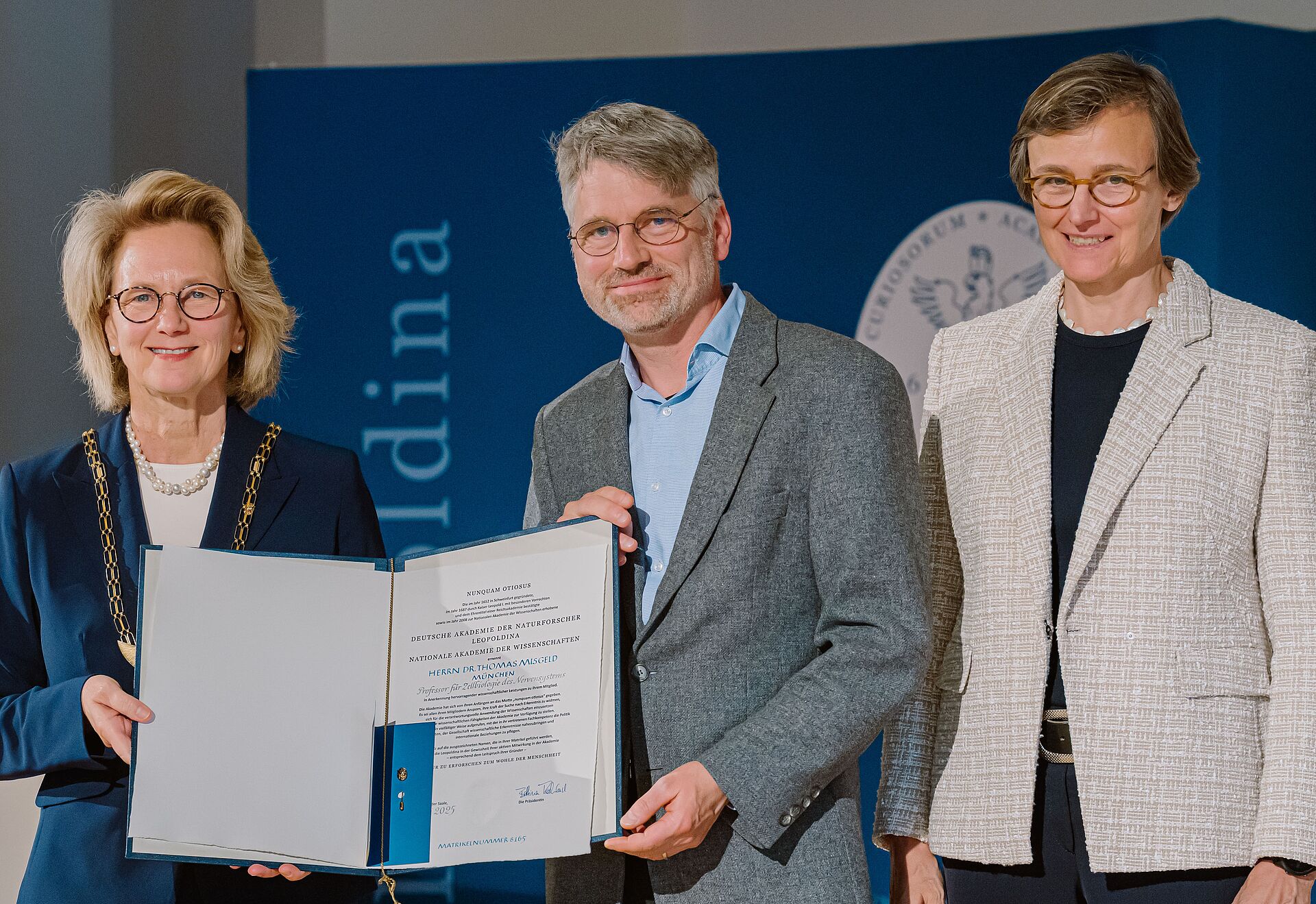 Three people stand on a Leopoldina stage presenting an open certificate during a scientific award or recognition ceremony.