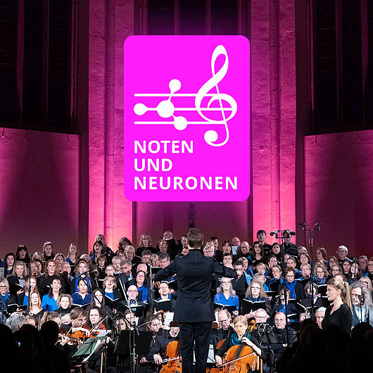 Das Foto zeigt ein großes Chor- und Orchesterensemble während eines Konzerts in einer Kirche oder Konzerthalle. Im Mittelpunkt steht eine dirigierende Person mit dem Rücken zum Publikum. Über den Mitwirkenden ist das magentafarbene Logo „Noten und Neuronen“ gut sichtbar. Die Bühne ist in Pink und Violett beleuchtet, während Chor, Solistinnen und Solisten sowie das Orchester gemeinsam auftreten. Das Bild dokumentiert eine Veranstaltung, die Musik und Neurowissenschaften miteinander verbindet.