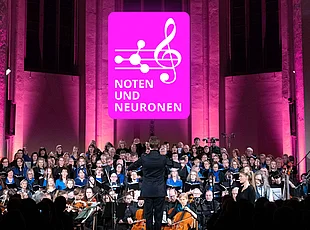 Das Foto zeigt ein großes Chor- und Orchesterensemble während eines Konzerts in einer Kirche oder Konzerthalle. Im Mittelpunkt steht eine dirigierende Person mit dem Rücken zum Publikum. Über den Mitwirkenden ist das magentafarbene Logo „Noten und Neuronen“ gut sichtbar. Die Bühne ist in Pink und Violett beleuchtet, während Chor, Solistinnen und Solisten sowie das Orchester gemeinsam auftreten. Das Bild dokumentiert eine Veranstaltung, die Musik und Neurowissenschaften miteinander verbindet.