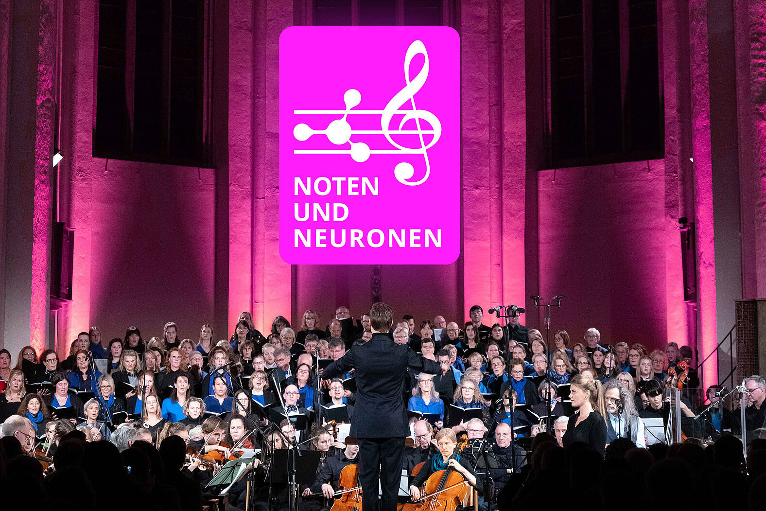 The photo shows a large choir and orchestra performing in a church or concert hall. A conductor stands at the center facing the performers with their back to the audience. Above the ensemble, a large magenta sign displays the event title “Notes and Neurons.” The stage is illuminated with pink and purple lighting while the choir, soloists, and orchestra perform together. The image documents a public event combining music and neuroscience.