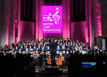 Das Foto zeigt ein großes Chor- und Orchesterensemble während eines Konzerts in einer Kirche oder Konzerthalle. Im Mittelpunkt steht eine dirigierende Person mit dem Rücken zum Publikum. Über den Mitwirkenden ist das magentafarbene Logo „Noten und Neuronen“ gut sichtbar. Die Bühne ist in Pink und Violett beleuchtet, während Chor, Solistinnen und Solisten sowie das Orchester gemeinsam auftreten. Das Bild dokumentiert eine Veranstaltung, die Musik und Neurowissenschaften miteinander verbindet.