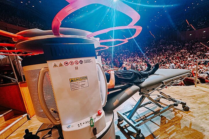 A person lies in a mobile MRI scanner on a stage, with a full concert hall visible in the background.