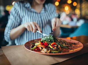 Das Bild zeigt eine Frau beim Essen in einem Restaurant. Im Vordergrund steht ein Teller mit gegrilltem Gemüse, darunter Paprika, Zucchini und Pilze, sowie grünem Salat. Die Frau hält Messer und Gabel in den Händen, während der Restaurantbereich im Hintergrund unscharf zu erkennen ist.