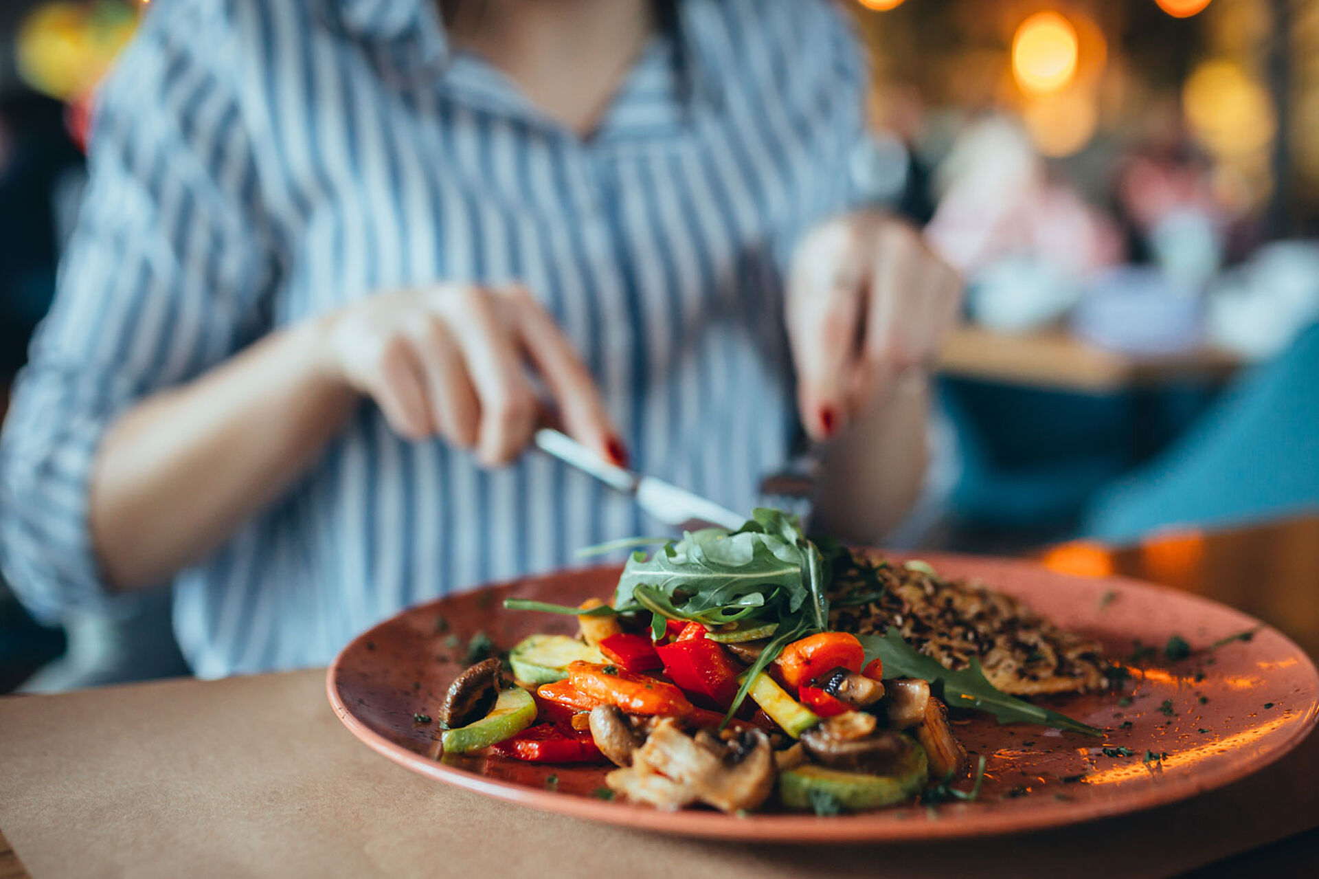 Das Bild zeigt eine Frau beim Essen in einem Restaurant. Im Vordergrund steht ein Teller mit gegrilltem Gemüse, darunter Paprika, Zucchini und Pilze, sowie grünem Salat. Die Frau hält Messer und Gabel in den Händen, während der Restaurantbereich im Hintergrund unscharf zu erkennen ist.