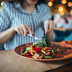 The image shows a woman eating at a restaurant. In the foreground is a plate of grilled vegetables, including peppers, zucchini, and mushrooms, topped with green salad. The woman holds a knife and fork, while the restaurant interior is blurred in the background.