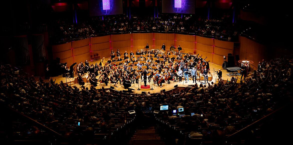 Das Bild zeigt einen großen Konzertsaal aus der Vogelperspektive. Auf der Bühne musizieren ein Sinfonieorchester sowie weitere Mitwirkende vor einem voll besetzten Publikum. Über der Bühne sind zwei große Leinwände mit dem Logo der Veranstaltungsreihe „Noten und Neuronen“ zu sehen. Die Aufnahme vermittelt die Größe und Atmosphäre der Veranstaltung, bei der Musik und Neurowissenschaften zusammengeführt werden.