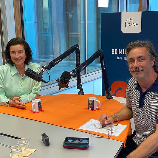Dorothee Bär und Moderator Marco Schreyl sitzen sich bei einer Aufnahme des DZNE-Hirnpodcasts „90 Milliarden“ gegenüber. Beide blicken lächelnd in die Kamera. Auf dem Tisch befinden sich zwei Studiomikrofone, im Hintergrund ist ein DZNE-Aufsteller zum Podcast zu sehen.