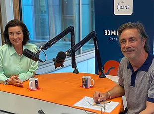 Dorothee Bär und Moderator Marco Schreyl sitzen sich bei einer Aufnahme des DZNE-Hirnpodcasts „90 Milliarden“ gegenüber. Beide blicken lächelnd in die Kamera. Auf dem Tisch befinden sich zwei Studiomikrofone, im Hintergrund ist ein DZNE-Aufsteller zum Podcast zu sehen.