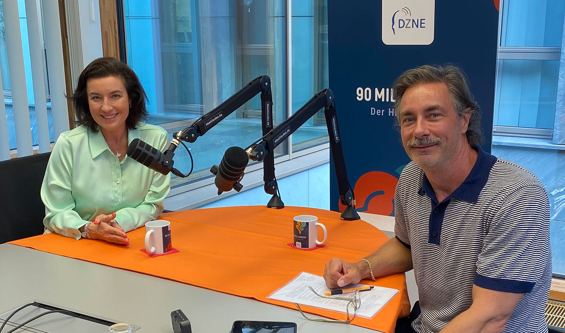 Dorothee Bär und Moderator Marco Schreyl sitzen sich bei einer Aufnahme des DZNE-Hirnpodcasts „90 Milliarden“ gegenüber. Beide blicken lächelnd in die Kamera. Auf dem Tisch befinden sich zwei Studiomikrofone, im Hintergrund ist ein DZNE-Aufsteller zum Podcast zu sehen.
