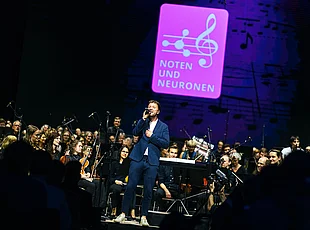 Ein Sänger im blauen Anzug steht mit einem Mikrofon vor einem Orchester auf der Bühne. Im Hintergrund leuchtet das Logo der Veranstaltung „Noten und Neuronen“.