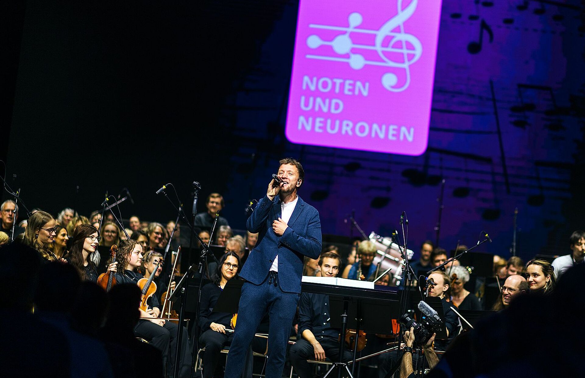 Ein Sänger im blauen Anzug steht mit einem Mikrofon vor einem Orchester auf der Bühne. Im Hintergrund leuchtet das Logo der Veranstaltung „Noten und Neuronen“.